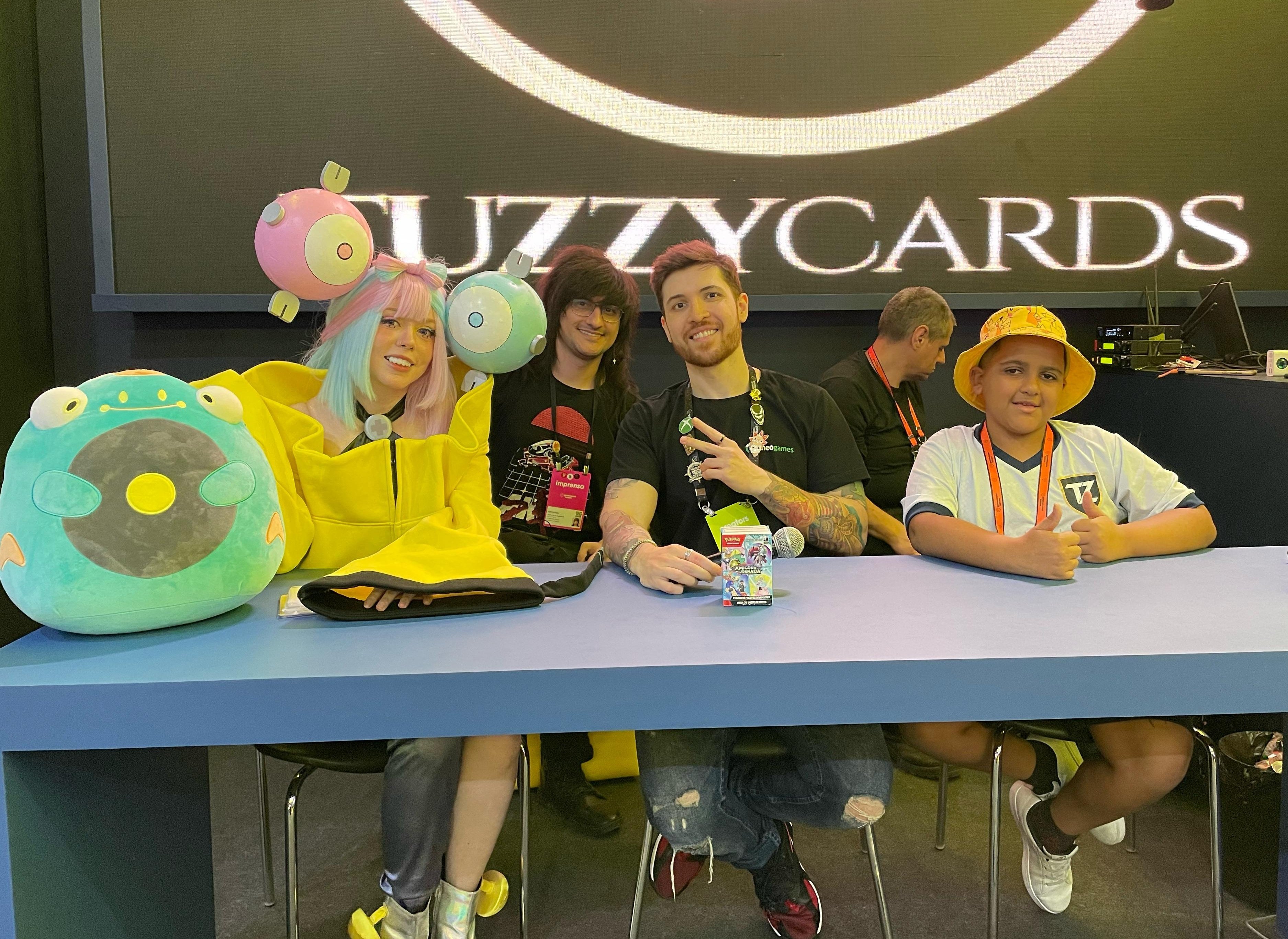 Foto do Palco Tuzzy Cards, com a presença de feekachips de cosplay de Kissara (Fernanda) à esquerda, Fábio Celestino e KingVenom (Marcelo) ao centro, e campeão Kauan à direita